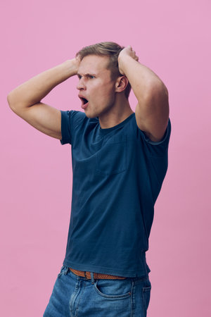 Young man with short hair wears casual blue t-shirt, expressing surprise and shock, holding his head...