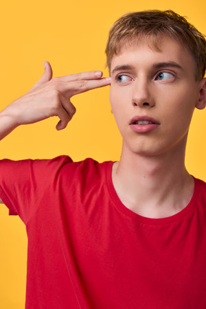 Gesture of a thoughtful youth with finger toward temple, red shirt against a bright yellow backdrop, conveying curiosity, hesitation, or playful thinking in a modern styleの写真素材