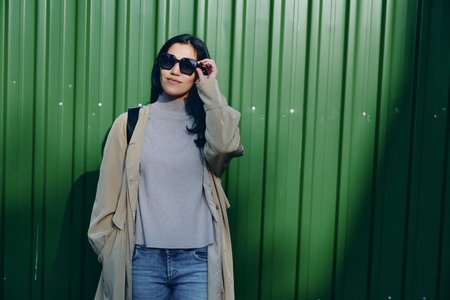 Woman wearing sunglasses and a beige coat stands against a green wall, casual fashion style in an outdoor scene, showcasing modern outfit details and confident demeanorの写真素材