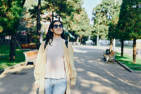 Street fashion of a woman in a light coat and jeans walking through a sunlit park, sunglasses on, relaxed stride, urban nature setting, fashionable outdoor moment ideal for lifestyle and travelの写真素材