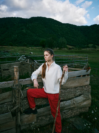 Woman on a rustic fence in a green countryside, holding a wooden staff, wearing white sweater and red pants, with hills in the background and cloudy sky.の写真素材