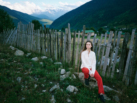 Woman outdoor fence mountains valley landscape, sits beside a weathered wooden fence in a lush valley with distant mountains, wearing red pants and a light top, capturing a calm outdoor moment inの写真素材