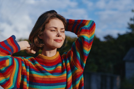 Woman in rainbow sweater smiling with eyes closed, young female model leaning back with hands behind head outdoors in sunlit park, relaxed cozy knit portrait for lifestyle fashion.の写真素材