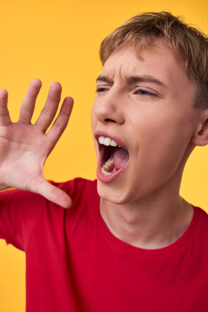 Young man shouting into his cupped hand on a bright yellow background, wearing a red shirt,...