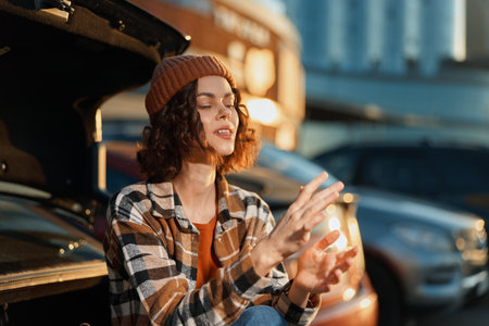 woman car trunk beanie plaid urban relaxation candid young woman sits at open car trunk during golden hour, eyes closed with hands warm in cozy beanie, mindful living and emotional storytellingの写真素材