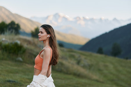 A serene woman stands in a grassy landscape with distant mountains under a clear blue sky. She wears...