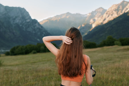 A woman with long hair stands in a grassy field, viewed from behind, as distant mountains rise. She...