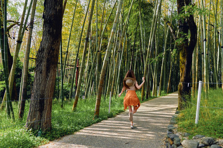 Woman in orange dress and straw hat walking along a gravel path through tall bamboo forest, summer sunlight filtering through green leaves, carefree motion on a nature trail.の写真素材