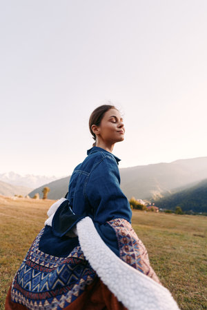 Woman portrait in nature with mountains and meadow, sunlight on her face as she breathes fresh air, eyes closed and relaxed wearing a denim jacket and patterned skirt at golden hour.の写真素材