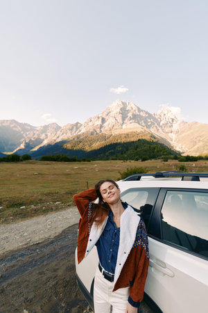 Woman by car in mountains and nature, travel portrait on road and meadow. Young woman leaning on SUV, relaxed pose, scenic alpine landscape and sunny outdoor adventure.の写真素材