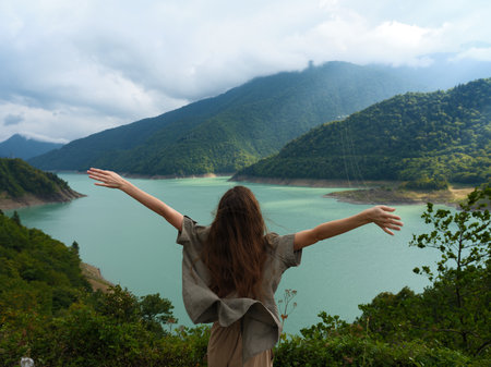 Woman with arms outstretched overlooking turquoise lake and mountains, nature landscape scene with forest foreground, sense of freedom, travel, solitude and outdoor exploration.の写真素材