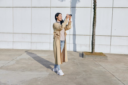 Selfie moment: a woman captures a sunny outdoor scene in a beige coat, denim jeans and white sneakers, standing beside a palm tree against a clean wall, sharing candid fashion and daily life throughの写真素材