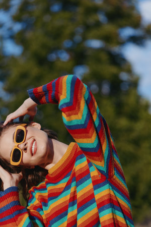 Woman smiling in striped sweater and sunglasses outdoor. Young adult model in casual outfit leaning back with joyful expression, colorful fashion portrait for lifestyle and travel use.の写真素材