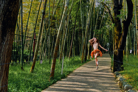 Bamboo forest, woman running on path in nature, joyful girl in orange dress skipping along sunlit walkway among tall bamboo trees, motion, freedom and outdoor leisure.の写真素材