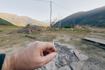 Hand holding cigarette butt above cold campfire ashes in rural mountains near a wooden bench and grassy field, outdoor camping scene with late afternoon light, solitude and quiet nature.の写真素材