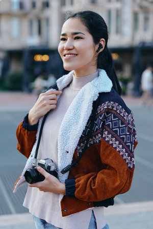 Smiling woman outdoors with a camera, wearing a patterned jacket, capturing city life in a candid photography moment, casual style, travel vibe, and creative street sceneの写真素材