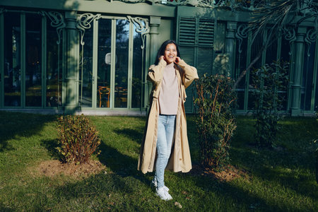 Woman wearing a long beige coat outdoors in a sunny garden, standing on grass near a glass conservatory, casual fashion and lifestyle moment in a peaceful suburban settingの写真素材