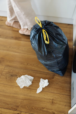 Garbage trash waste bag floor tissue near kitchen cabinet on hardwood, black tied plastic sack with yellow tie and crumpled paper on laminate, feet of person in background during home cleaning.の写真素材