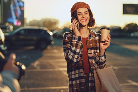 Woman holding coffee and shopping bag, smiling on phone in parking lot at sunset, candid lifestyle portrait with authenticity, golden hour glow, mindful living and emotional storytelling.の写真素材