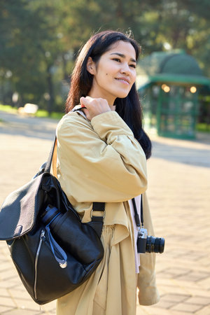 Young photographer in a park wearing a beige trench, bag over shoulder with camera, warm sunlight, exploring outdoor scenes for creative street photography and natural light shotsの写真素材