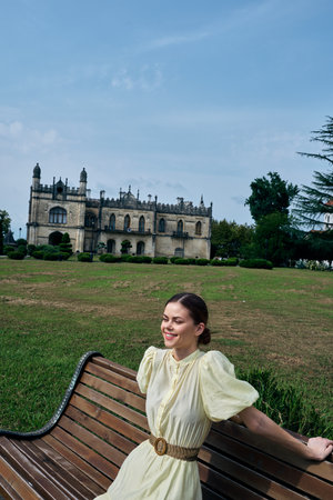 Woman in a pale dress sits on a bench in a green park with a historic castle in the background, smiling and enjoying a peaceful outdoor moment, on a sunny day, enjoying the scenery and calm ambiance.の写真素材