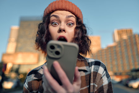 Woman holding phone with surprised expression in city street, candid authenticity captured in golden glow of sunset, emotional storytelling and mindful living urban moment.の写真素材