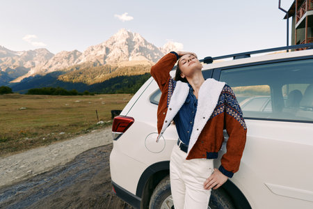 Woman leaning on car by mountains during travel, enjoying nature and open road outdoors, relaxed pose in cozy sweater, lifestyle vacation moment with scenic alpine backdrop.の写真素材