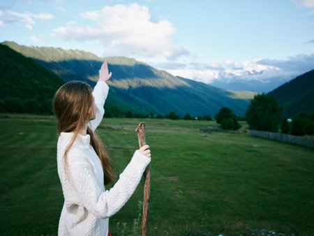 Outdoor scene with a girl in a white sweater raising a wooden stick toward distant mountains across...