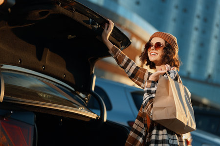 Woman shopping by car trunk with smile and bag in urban sunlight, candid lifestyle moment with...