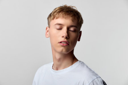 Young man with closed eyes wearing a white tshirt in a calm studio setting, a serene fashion pose that conveys relaxed mood, minimal light and quiet confidence in a simple candid styleの写真素材