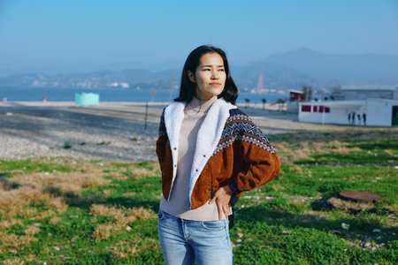 Woman in a warm jacket stands outdoors in a coastal meadow, hands on hips, bright daylight, casual...