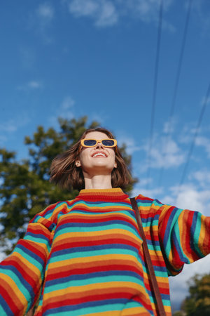 Young woman smiling in a colorful striped sweater and sunglasses outdoors under a bright blue sky,...