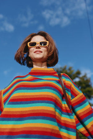 Woman in a colorful striped sweater and yellow sunglasses smiles confidently outdoors against a blue sky. Young adult with bob haircut and casual outfit, relaxed travel lifestyle portrait.の写真素材
