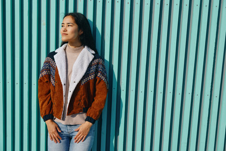 Woman in a warm shearling jacket stands against a turquoise striped wall showcasing casual...