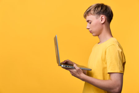 Boy holding a laptop in a bright yellow studio, focused on screen for learning, technology, and...