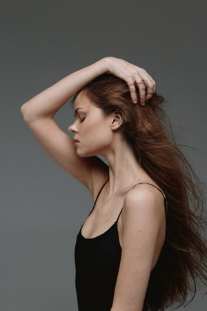thoughtful woman with curly hair in black top posing against gray background, calm and elegant...