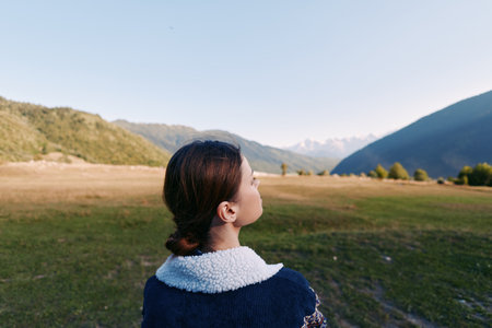 Woman with back to camera in mountain landscape and valley field, surrounded by nature and open...