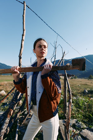 Woman holding axe on shoulder by wooden fence in rural countryside portrait, standing outdoors with...