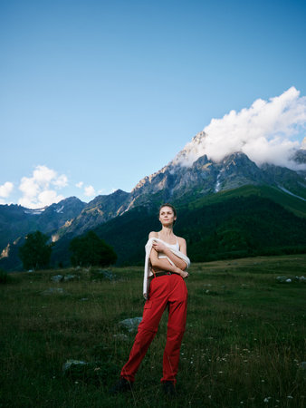 Mountain landscape with a wide meadow and blue sky, a woman in red pants stands confidently,...