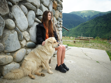 Girl sitting with a large dog beside a stone wall in a mountain landscape, outdoors, enjoying...