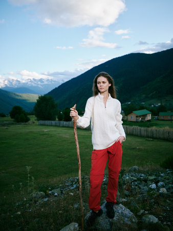 Outdoor scene of a woman standing on a rocky overlook with a wooden staff, mountains in the...