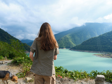 Woman stands with back to camera by turquoise lake and mountains, dog nearby on rocky shore, nature...