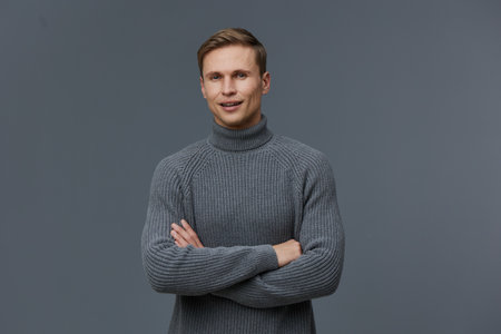 smiling young caucasian man in gray sweater stand pose crossed arms look camera isolated on gray...