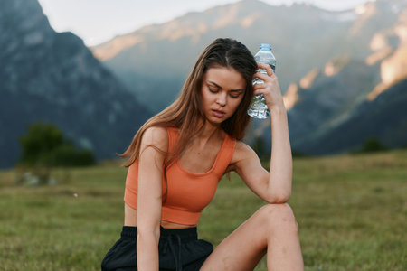 A young woman sits outdoors in a scenic valley with mountains in the background. She wears an orange tank top, dark shorts, and holds a water bottle, looking thoughtful while enjoying a peacefulの写真素材