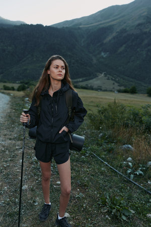 A young woman stands on a rugged trail in a mountain landscape, holding a trekking pole and camera...