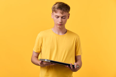 Young man in a yellow t shirt holds a tablet, concentrating on the screen in a bright studio, illustrating digital learning, online reading, and casual tech use.の写真素材
