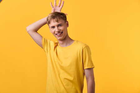 Playful man in a yellow shirt raises a hand against a bright yellow backdrop, conveying joy and casual energy for lifestyle, fashion, and positive mood imageryの写真素材