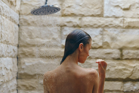 Woman showering outdoors under rain showerhead against natural stone wall, enjoying refreshing water...