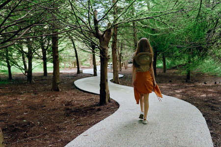 Woman walking on winding path through forest with backpack and orange dress, trail surrounded by...