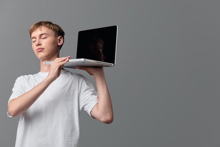 Young man balancing a laptop on his shoulder eyes closed calm expression casual white t shirt minimal studio setting conceptual moment of portable tech and focusの写真素材
